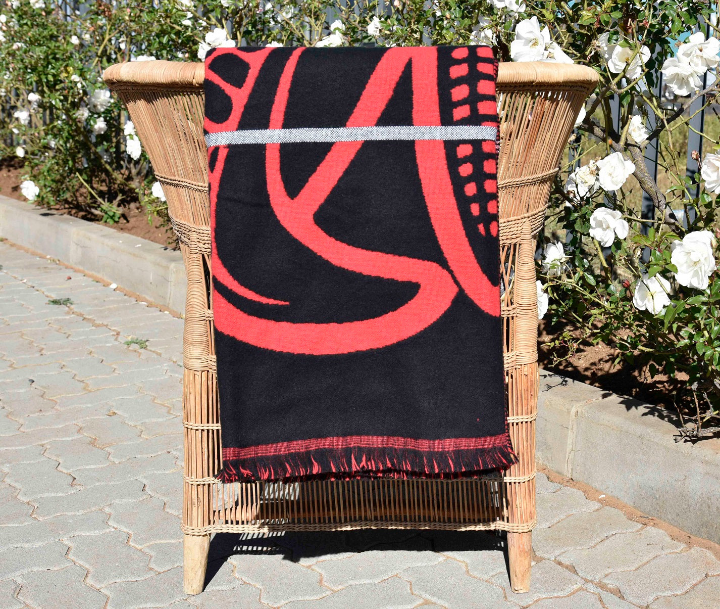 Black Basotho blanket with red abstract design draped over a wicker chair outdoors.
