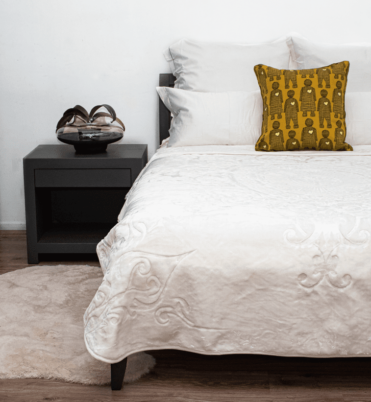 A styled bedroom scene showing a bed with white blanket over a bed and a mustard yellow decorative pillow featuring a black geometric pattern. A black nightstand with a pair of shoes sits beside the bed on a light rug against a white wall.
