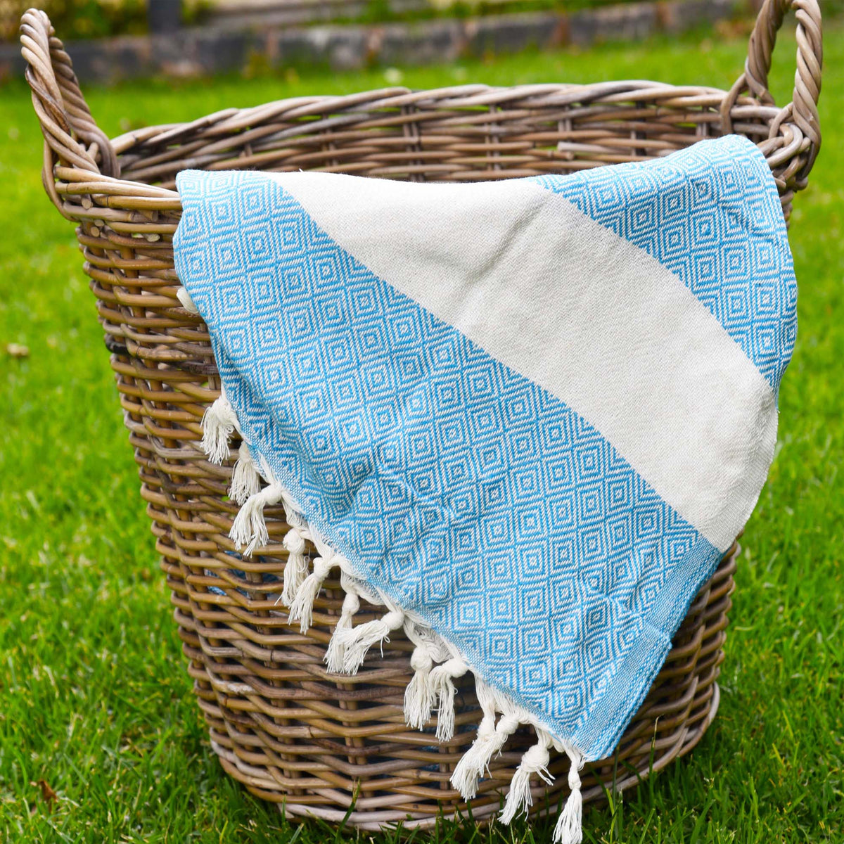 A turquoise and white patterned Turkish cotton towel with white tassels, draped over a rustic wicker basket on a green grass lawn.