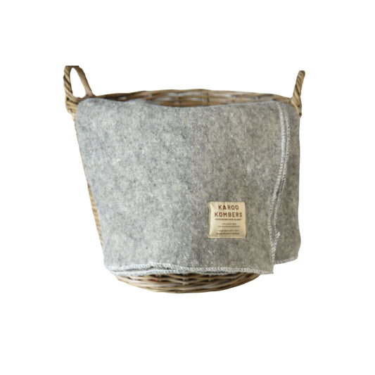 A grey wool blanket in a natural wicker basket, featuring a beige brand label that reads 'KAROO KOMBERS'