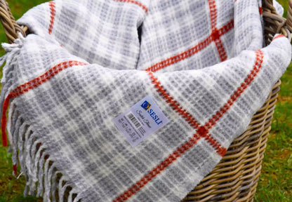 A close-up of a checkered scotch blanket with a white and grey plaid pattern and red stripes, draped over a wicker basket on grass.
