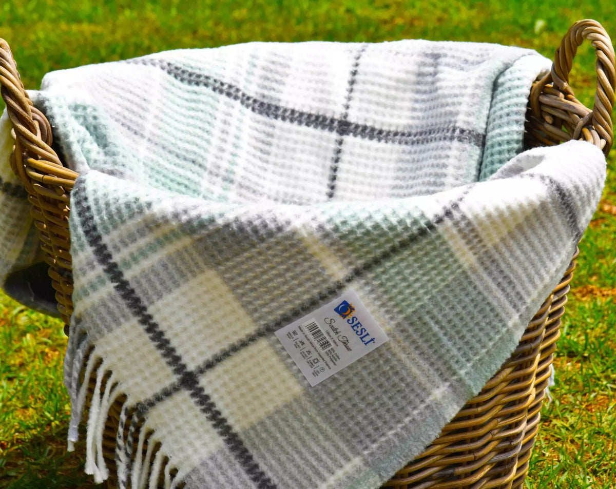 A plaid knit blanket with white, grey, and pale green stripes draped over a wicker basket on a grassy lawn. 