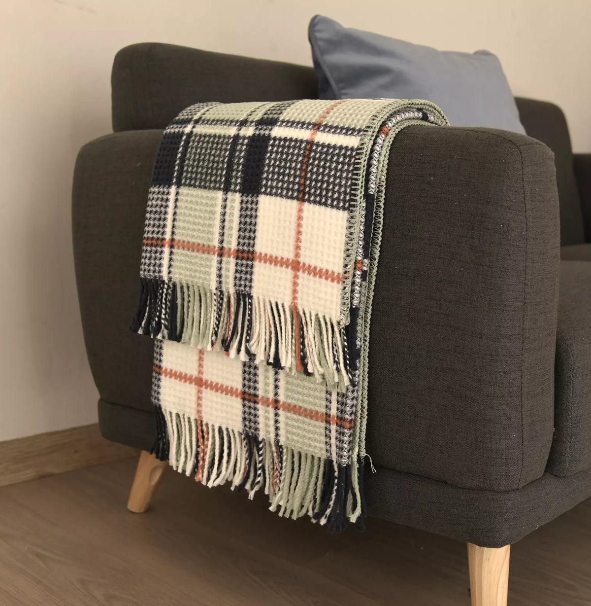 A woven plaid throw blanket with fringe, draped over the arm of a dark gray armchair in a living room setting.
