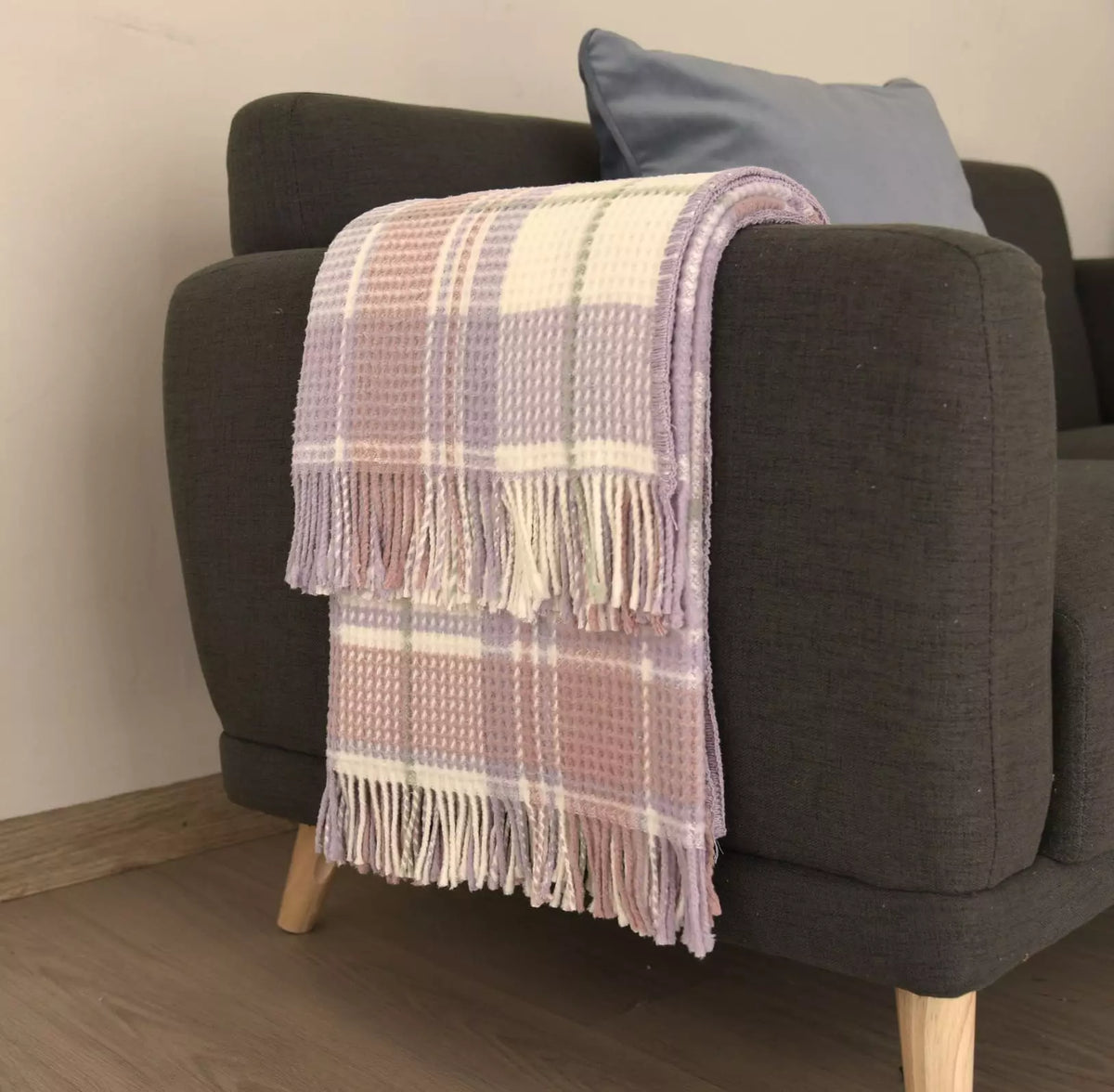 A pastel plaid throw blanket with fringe is draped over the arm of a dark gray armchair in a living room setting.