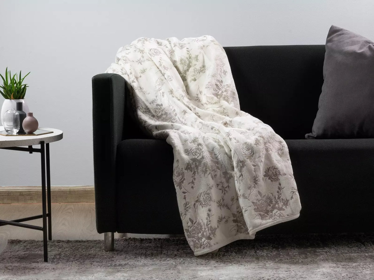 A cream-colored throw blanket with a muted gray floral toile pattern draped over a black sofa in a modern living room setting.