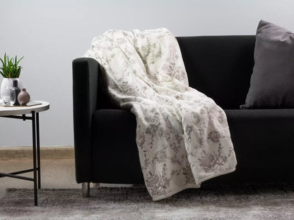 A cream-colored throw blanket with a muted gray floral toile pattern draped over a black sofa in a modern living room setting.