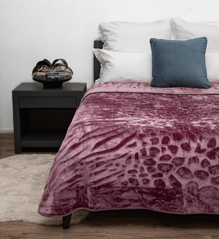 A plush, textured mauve-purple blanket with a raised organic pattern is draped over a bed in a bedroom setting, styled with white pillows, a slate blue accent pillow, a black nightstand, and a light wood floor.