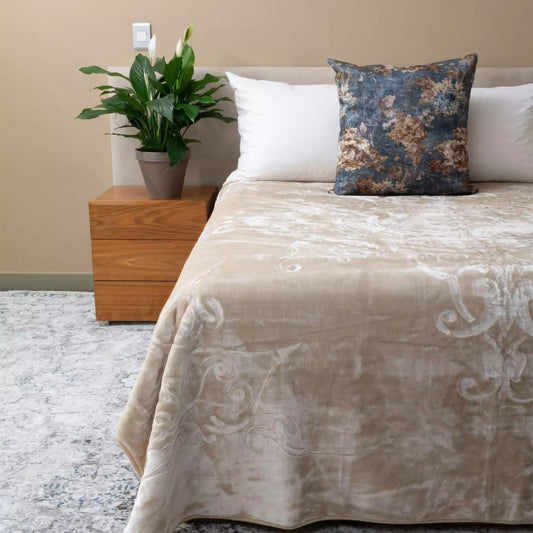 A beige velvet throw blanket with a subtle damask pattern is draped over a bed, styled with white pillows and a blue floral accent pillow. The bed is set in a bedroom with a wooden nightstand holding a potted green plant, a beige wall, and a gray patterned rug.