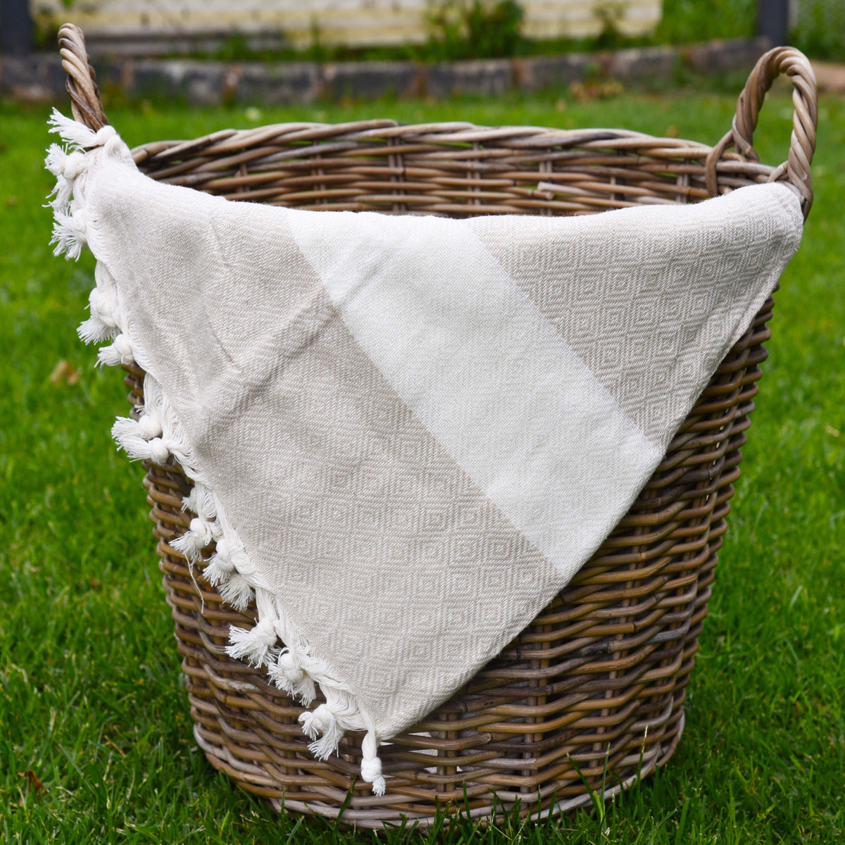A beige peshtemal towel with small diamond geometric shapes, a white stripe across the towel and frills at the bottom. the towel is placed in a woven basket sitting on grass. perfect for picnics and at the pool.