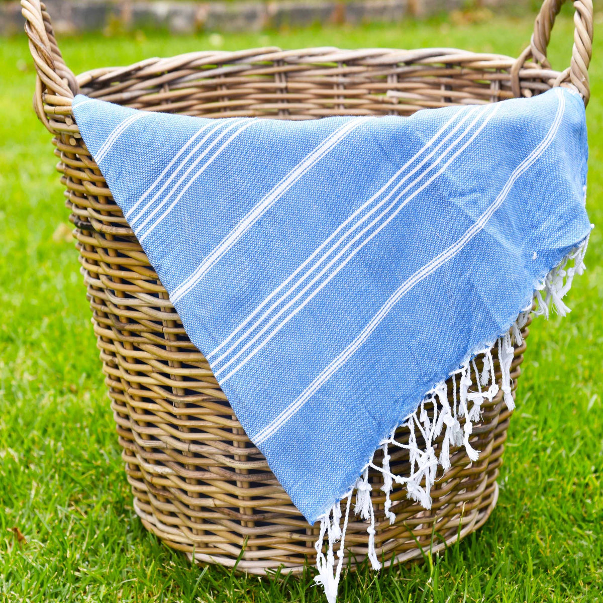 A light blue Turkish cotton towel with white stripes and fringed edges, draped over a natural wicker basket on a green grass lawn.