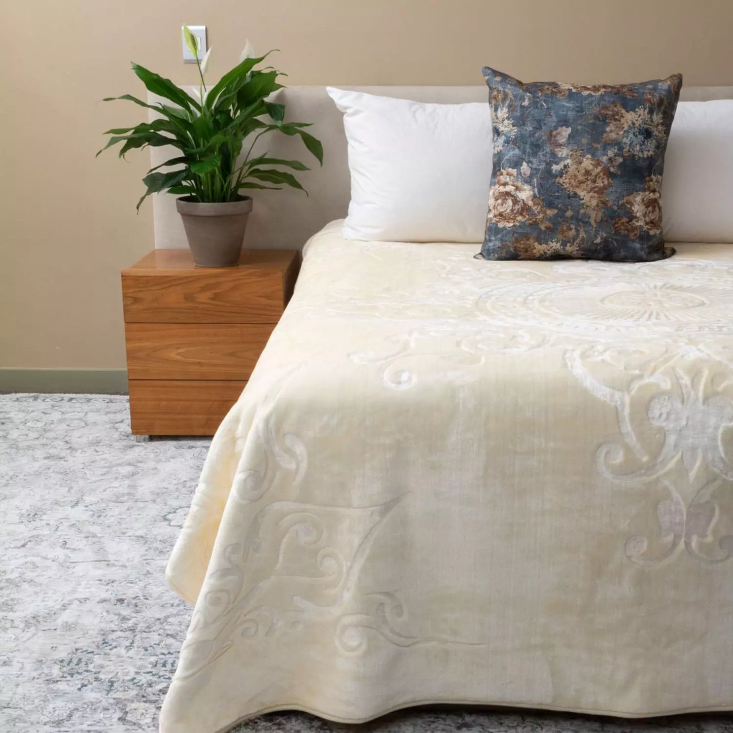 A cream-coloured textured blanket with a subtle damask pattern, styled on a bed with white pillows and a blue floral accent pillow, next to a wooden nightstand with a potted plant.