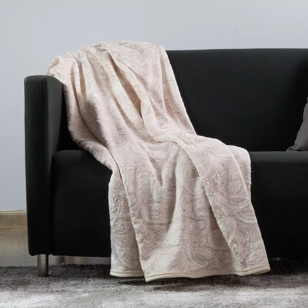 A dusty pink textured throw blanket with an intricate floral damask pattern draped over a modern black fabric sofa.