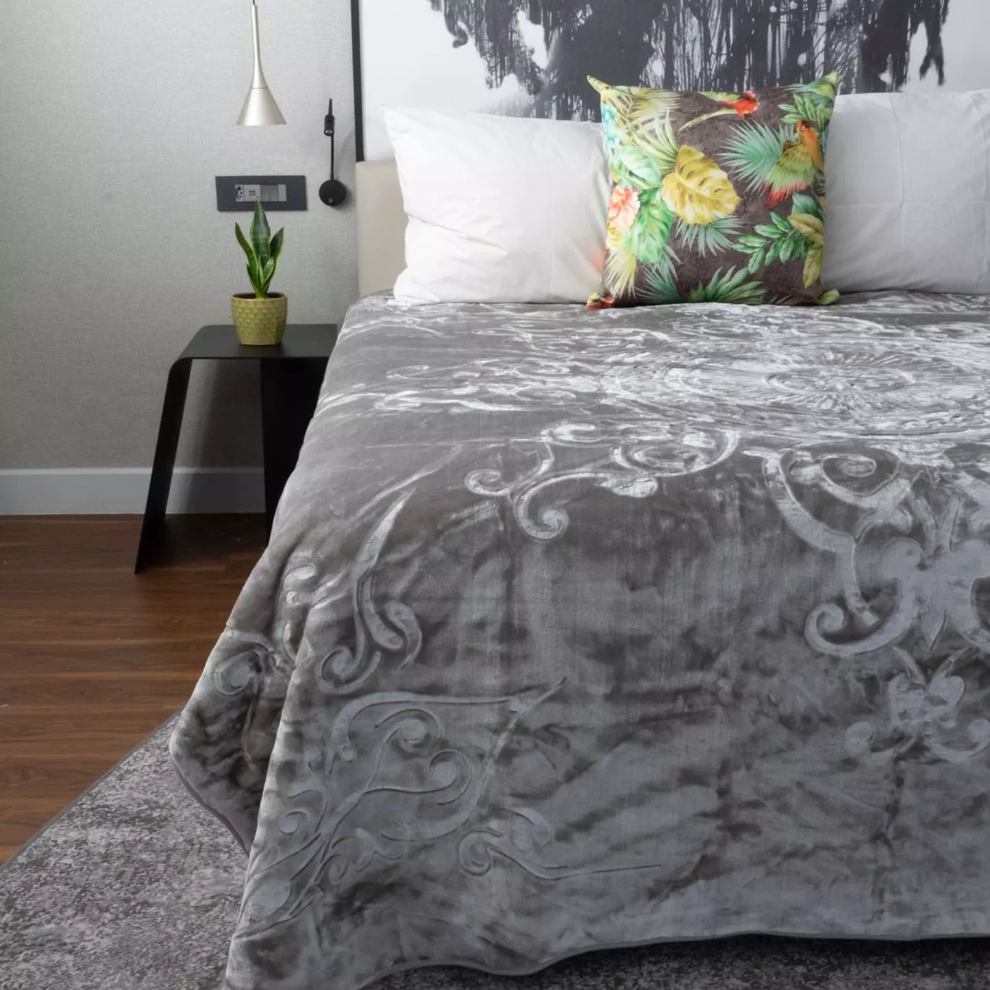 A gray blanket with a white baroque scroll pattern is draped over a bed in a modern bedroom setting, styled with white pillows and a green tropical print accent pillow.