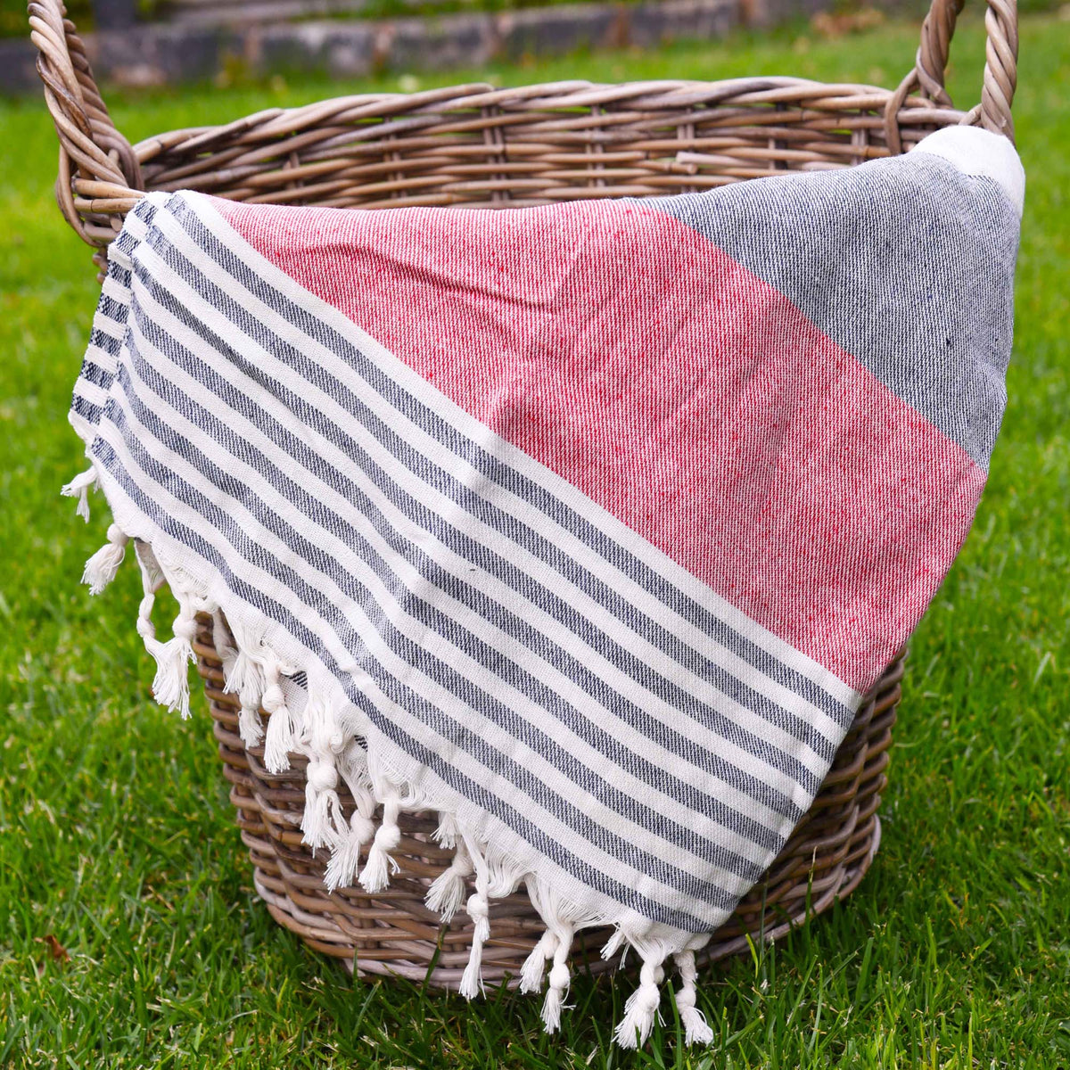 A woven Turkish towel with a color-blocked design in red, navy, and white stripes, draped over a wicker basket on a green grass lawn.