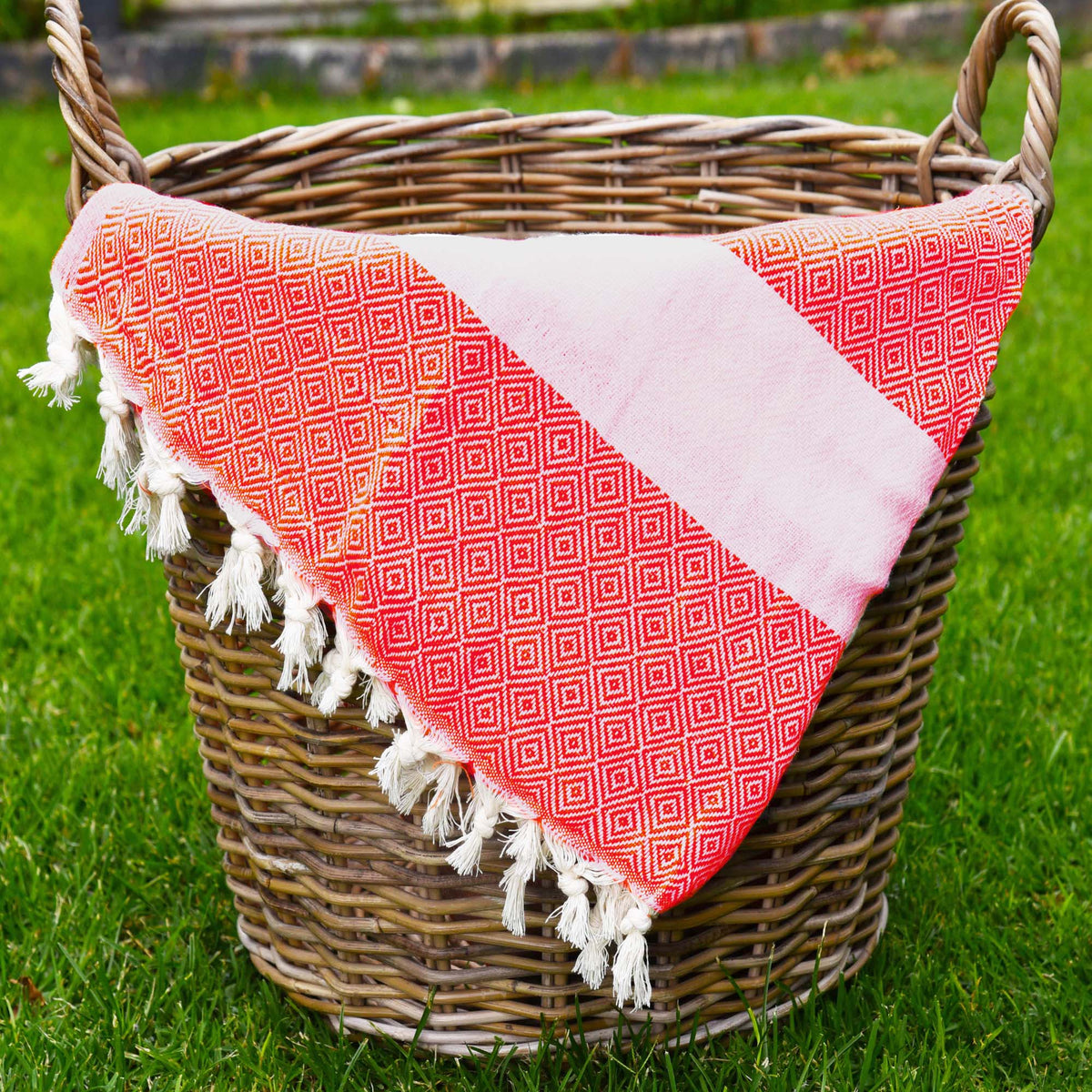 A red peshtemal towel with small diamond geometric shapes, a white stripe across the towel and frills at the bottom. The towel is placed in a woven basket sitting on the grass. Perfect for picnics and at the pool.