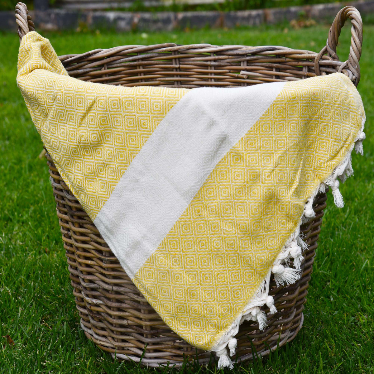 A yellow peshtemal towel with small diamond geormetric shapes, a white stripe across the towel and frills at the bottom. The towel is placed in a woven basket sitting on grass. perfect for picnics and pool side